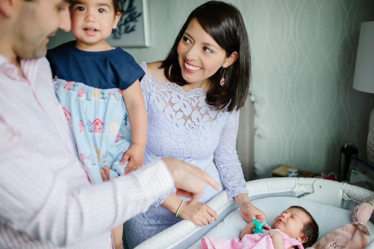 A mother smiling at her toddler beside a baby lying in a bassinet